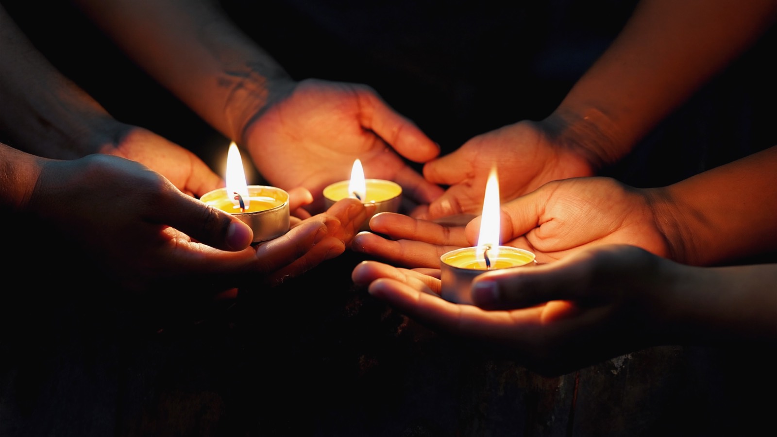 Mehrere Hände schützen kleine Kerzenflammen in der Dunkelheit, warme Lichtpunkte im schwarzen Umraum.